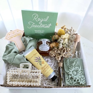May include: A gift box filled with self-care items. Includes a green tea box with the text "Royal Treatment", a jar of honey, a bouquet of dried flowers, a tube of honey lavender hand cream, a pearl hair clip, a green scrunchie, and a floral soap.