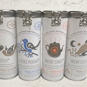 Op de afbeelding: Vier zilveren theeblikken met kleurrijke illustraties en tekst. De blikken zijn gelabeld met "Chokola", "Bluebird Morning", "Ancient Sunrise" en "Dream Catcher". Elk blik heeft een ander vogelontwerp en bevat de tekst "Flying Bird Botanicals" en "100% Organic Tea".