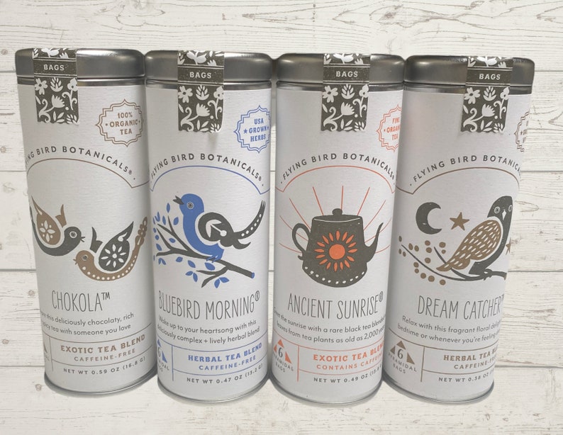 Op de afbeelding: Vier zilveren theeblikken met kleurrijke illustraties en tekst. De blikken zijn gelabeld met "Chokola", "Bluebird Morning", "Ancient Sunrise" en "Dream Catcher". Elk blik heeft een ander vogelontwerp en bevat de tekst "Flying Bird Botanicals" en "100% Organic Tea".
