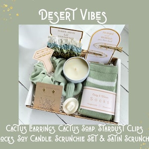 May include: A gift box with the text "Desert Vibes" contains a variety of items. These include a scrunchie set, socks, a soy candle, cactus earrings, cactus soap, and hair clips. The box is decorated with a desert theme.