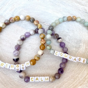 Könnte beinhalten: Drei Armbänder mit Perlen mit den Wörtern "STRENGTH", "CALM" und "COURAGE" in weißen Buchstabenperlen. Die Armbänder weisen eine Mischung aus bunten, runden Edelsteinperlen in Lila-, Braun- und Grüntönen auf.