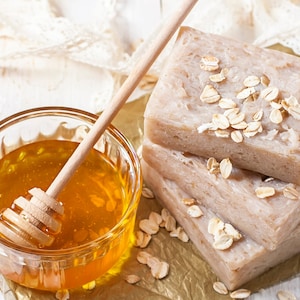 May include: A close-up of a glass bowl filled with honey and a wooden honey dipper. Three bars of oatmeal soap are stacked on top of each other. The soap is decorated with oatmeal flakes.