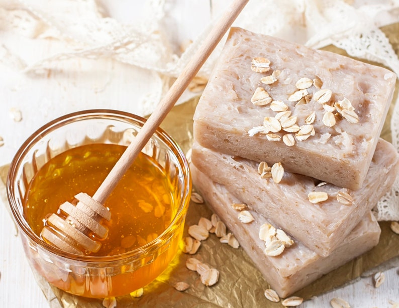 May include: A close-up of a glass bowl filled with honey and a wooden honey dipper. Three bars of oatmeal soap are stacked on top of each other. The soap is decorated with oatmeal flakes.
