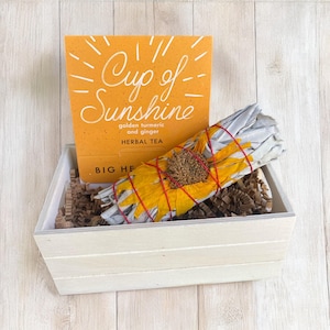 May include: A white wooden box filled with brown packing peanuts and a white sage smudge stick wrapped with a dried sunflower. A yellow card with the text "Cup of Sunshine golden turmeric and ginger HERBAL TEA BIG HE" is visible in the background.