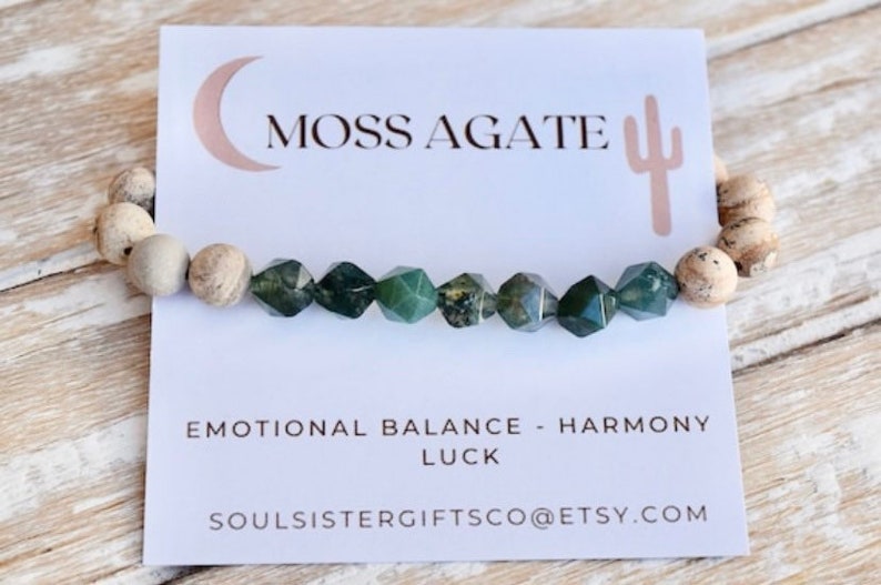 May include: A green moss agate bracelet with white and brown jasper beads. The bracelet is on a white card with the text "Moss Agate", "Emotional Balance - Harmony", "Luck", and "Soulsistergiftsco@etsy.com".