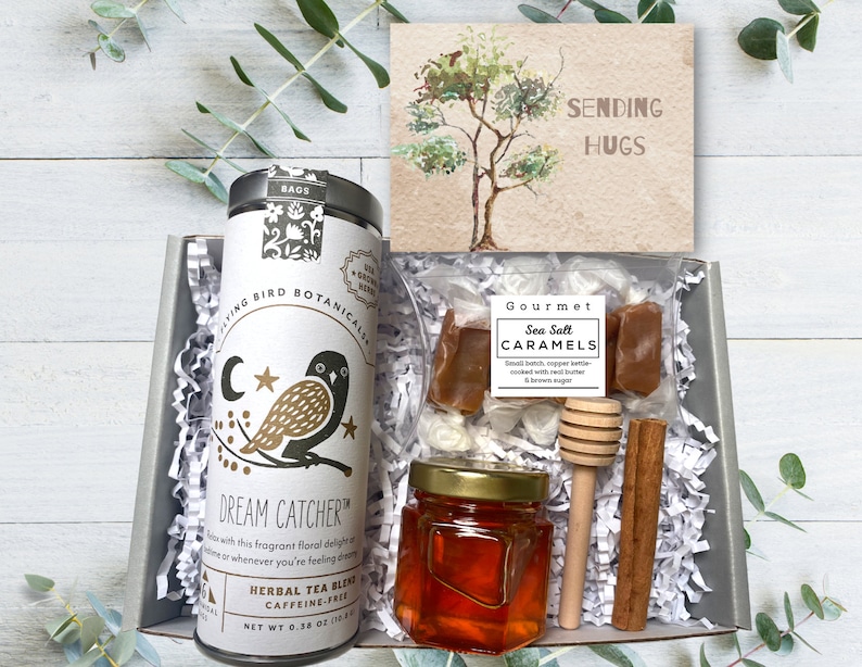 Op de afbeelding: Een cadeaudoos gevuld met verschillende items, waaronder een blikje kruidenthee, een pot honing, een zak zeezoutkaramellen, een houten honingroerder, een kaneelstokje en een kaart met de tekst "Sending Hugs".