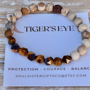 May include: A brown and gold tiger's eye gemstone bracelet with white and brown beads. The bracelet is on a white card with the text "Tiger's Eye", "Protection - Courage - Balance", and "Soulsistergiftsco@etsy.com".
