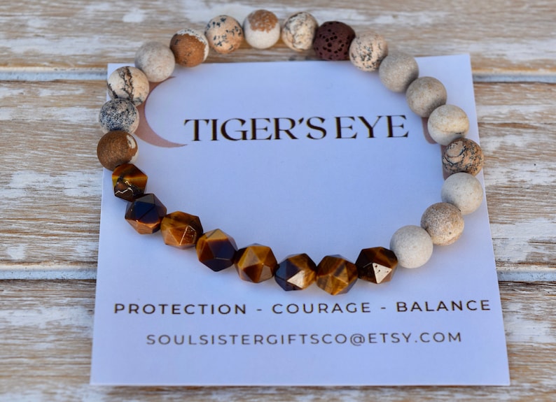 May include: A brown and gold tiger's eye gemstone bracelet with white and brown beads. The bracelet is on a white card with the text "Tiger's Eye", "Protection - Courage - Balance", and "Soulsistergiftsco@etsy.com".