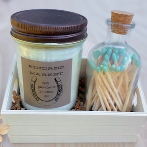 Peut inclure: Une boîte en bois blanche contenant une bougie en soja et une bouteille en verre remplie d'allumettes. Le pot de bougie a une étiquette brune avec le texte "KINDRED MARKET" et "100% Hand-Crafted Soy Candle 8oz". Les allumettes ont des pointes bleues.