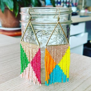May include: Two pairs of triangle-shaped earrings with gold hoops and colorful beaded fringe. One pair has green, pink, and white fringe. The other pair has orange, blue, and yellow fringe.