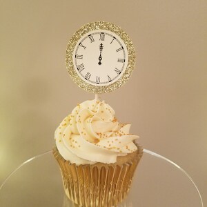 May include: A cupcake with white frosting and gold sprinkles, topped with a clock-shaped decoration. The clock shows the time as midnight, with a gold glitter border. The cupcake sits in a gold foil wrapper.
