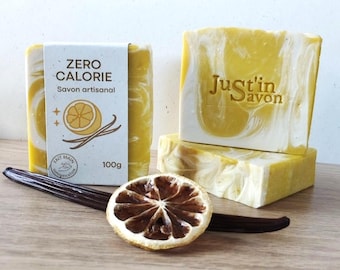 Può includere: Barrette di sapone artigianali con un design a spirale giallo e bianco, etichettate "Zero Calorie" e "Just'in Savon". L'immagine include una fetta di limone essiccata e una bacca di vaniglia. Ogni barretta pesa 100g.