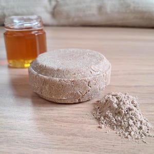 Peut inclure: Un pain de shampooing rond brun clair, à côté d'une petite pile de poudre et d'un pot de miel. Le pain de shampooing a une surface texturée. Le miel est dans un pot en verre transparent.