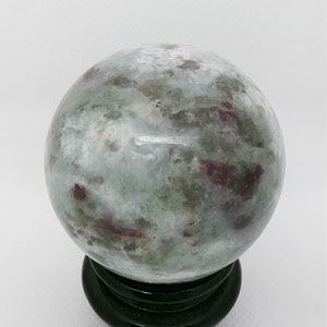 Peut inclure: Une sphère verte et blanche avec des taches rouges et roses, posée sur un support en bois noir. La sphère est une pierre polie.