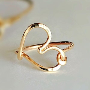 May include: A gold heart-shaped ring with a knot detail. The ring is made of wire and is a simple, delicate design.