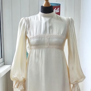 May include: A floor-length cream-colored dress featuring long bishop sleeves with ruffled cuffs, a high neckline, and an empire waist. The garment is accented with intricate white lace trim along the collar, bodice, and hemline, creating a structured silhouette on a mannequin.