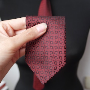 May include: A burgundy tie with a repeating geometric pattern in black. The tie is displayed on a black mannequin. The tie is folded in half, showcasing the intricate design. The tie is a classic accessory.