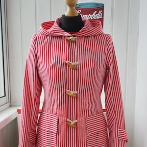 May include: A red and white striped hooded coat with wooden toggle closures and two front pockets. The coat is paired with a black and white patterned skirt. The outfit is displayed on a mannequin.