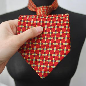 Może przedstawiać: Czerwony krawat z powtarzającym się wzorem żółtych i szarych kwadratów. Krawat jest trzymany przed czarnym manekinem. Węzeł krawata jest widoczny wokół szyi manekina.