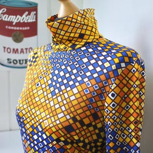 Könnte beinhalten: Ein langärmliges Rollkragenshirt mit einem geometrischen Muster aus gelben, orangefarbenen, blauen und weißen Quadraten. Das Kleidungsstück wird auf einer Schaufensterpuppe präsentiert. Eine Campbell's Tomatensuppendose ist im Hintergrund zu sehen.