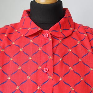 May include: A red button-down shirt with a repeating pattern of blue curved lines and yellow floral accents. The shirt has a rounded collar and is buttoned up. The shirt is displayed on a mannequin.