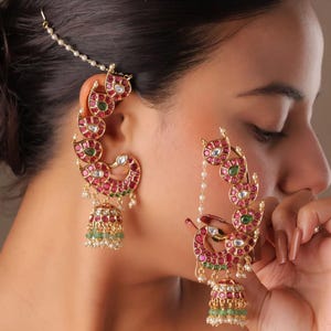 May include: Elaborate gold earrings with a peacock motif, adorned with red and green gemstones. The earrings have a dangling bell-shaped base with green beads and small white pearls. A delicate chain with pearls attaches to the hair.