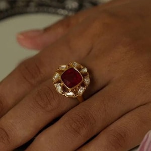 May include: A gold ring with a red gemstone surrounded by smaller clear gemstones. The ring is on a finger of a hand resting on a book.