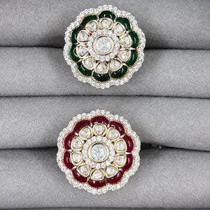 May include: Two floral-shaped rings with a central clear stone and a halo of smaller stones. One ring has a green enamel border, the other a burgundy enamel border. The rings are set on a gray surface.
