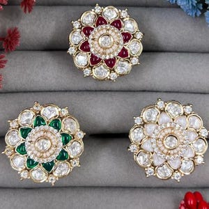以下が含まれることがあります： 宝石のアクセントが付いた、3つの豪華な金色の花のブローチ。1つのブローチは赤い中心部、もう1つは緑色、3つ目は白色です。各ブローチには中心に真珠があり、透明で輝く石で囲まれています。