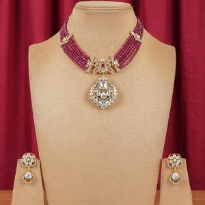 Puede incluir: Un collar de oro con un gran colgante adornado con piedras blancas. El collar está hecho de múltiples hilos de pequeñas cuentas rojas. También se incluyen pendientes a juego con piedras blancas.