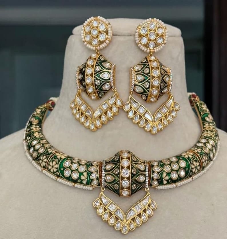 May include: A gold-toned jewelry set featuring a necklace and matching earrings. The necklace and earrings have a green enamel design with white floral accents and clear gemstones. The earrings are a drop style, and the necklace is a choker.