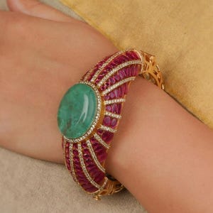 May include: A gold bracelet with a large, oval green gemstone. The bracelet is decorated with rows of small, red gemstones and clear accents. The design is intricate and ornate, showcasing a luxurious aesthetic. The bracelet is likely made of gold.