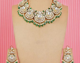 May include: A gold-toned jewelry set featuring a necklace, earrings, and a headpiece. The necklace and earrings have a floral design with white stones and green accents. The headpiece is a smaller version of the necklace. The set is displayed on a mannequin.