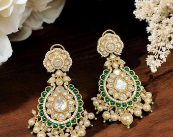 May include: A pair of gold-toned drop earrings with a floral design. The earrings feature clear, round gemstones, green accents, and dangling faux pearls. The earrings are set against a dark wooden surface.