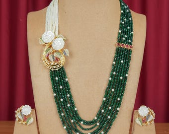 Könnte beinhalten: Eine mehrsträngige grüne Perlenkette mit weißen Perlenakzenten und einem dekorativen goldenen Anhänger mit weißen Blumenakzenten. Passende Ohrringe mit ähnlichen floralen Designs werden auf Ständern ausgestellt.