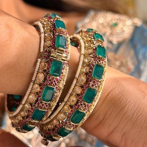 Pode incluir: Duas pulseiras douradas adornadas com pedras verdes lapidadas em esmeralda, pequenas pedras vermelhas e pérolas brancas. O design intrincado apresenta uma mistura de cores e texturas, criando uma aparência luxuosa.