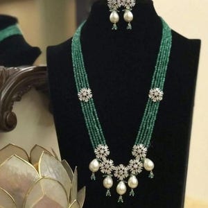 Puede incluir: Un collar de cuentas verdes y un juego de pendientes a juego exhibidos en un maniquí negro. El collar presenta múltiples hilos de cuentas verdes, acentuados con adornos en forma de flor y grandes perlas blancas en forma de lágrima. Los pendientes tienen un diseño similar.