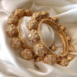 May include: Two gold bangles with intricate filigree and diamond-like embellishments. The bangles are adorned with multiple decorative, round elements. The jewelry rests on a white, silky fabric, creating a luxurious display.