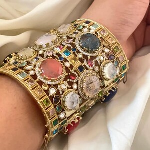 Pode incluir: Uma pulseira larga em tom dourado com um design de filigrana intrincado, incrustada com numerosas pedras preciosas cabochão multicoloridas, incluindo tons de rosa, azul, branco e vermelho, rodeadas por pequenos cristais claros em pavé.