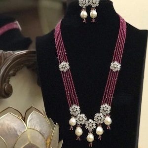 Puede incluir: Un collar de múltiples hilos con cuentas rosadas y un colgante con un racimo de piedras blancas y perlas. Pendientes a juego con piedras blancas y perlas.