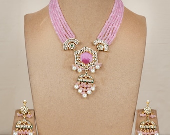 May include: A pink beaded necklace and matching earrings set. The necklace features a large, pink gemstone pendant with pearl and gold accents. The earrings have a similar design, with pink beads and pearl drops.