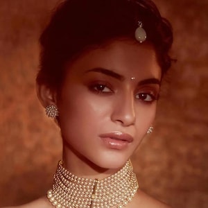 Peut inclure: Une femme porte un collier ras du cou doré à plusieurs rangs de perles, assorti de boucles d'oreilles et d'un petit bijou de tête. Les bijoux se détachent sur un fond chaud et neutre.
