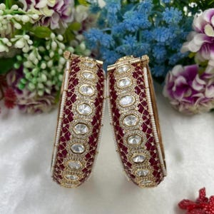 May include: Two ornate bangles with a gold-tone finish, featuring a deep red background and clear, oval-shaped gemstones. The bangles are further embellished with smaller white beads and intricate gold detailing, creating a luxurious look.