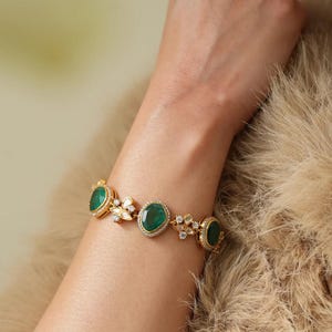 May include: A gold bracelet featuring oval-cut emeralds and small white gemstones. The bracelet has a delicate design with a series of emeralds set in gold, interspersed with floral-shaped accents. The bracelet is worn on a wrist.
