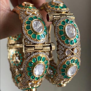 May include: Two gold bangles with intricate detailing. Each bangle features a central clear stone surrounded by green enamel and small clear stones. The bangles have a hinged clasp and are adorned with additional clear stones.