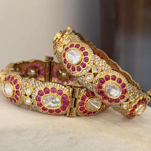 May include: Two gold-toned bangles with intricate detailing. Each bangle features a series of oval-shaped, clear stones surrounded by a ring of small, red stones. The bangles are further embellished with smaller, clear stones, creating a luxurious and ornate appearance. The bangles are likely for jewelry.