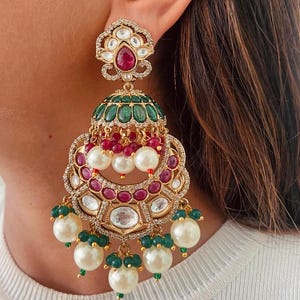 May include: Gold-toned chandelier earrings with a floral design. The earrings feature a large red gemstone at the top, green and red accents, and dangling pearls. The earrings are embellished with clear crystals.
