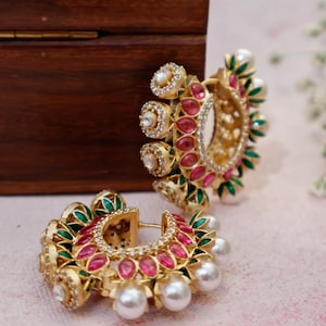 Puede incluir: Un par de pendientes de aro dorados. Cada pendiente presenta una forma de media luna adornada con piedras preciosas rojas y verdes, cristales transparentes y perlas blancas. Los pendientes se muestran sobre una superficie rosa.