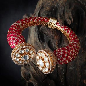 Op de afbeelding: Een goud-rode armband met een gedetailleerd ontwerp. De armband heeft een rode, patroonband en twee grote, ovale versieringen bezet met heldere stenen. De armband is geplaatst tegen een donkere, getextureerde achtergrond.