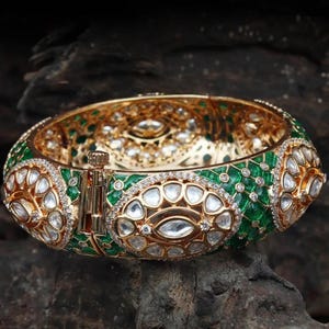 Könnte beinhalten: Ein goldfarbenes Armreif-Armband mit aufwendigen Details. Das Armband hat ein grünes Emaille-Design mit klaren, ovalen Steinen und kleineren klaren Steinen. Der Verschluss ist goldfarben, und das Armband liegt auf einem dunklen, strukturierten Hintergrund.